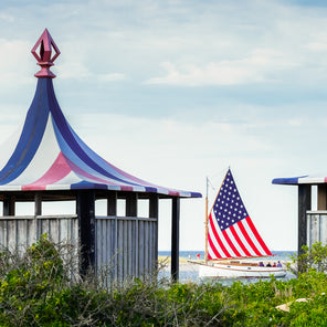 A panoramic photograph of red, white, and blue tented huts in Edgartown, Martha's Vineyard with a lighthouse and sailboat in the distance. 