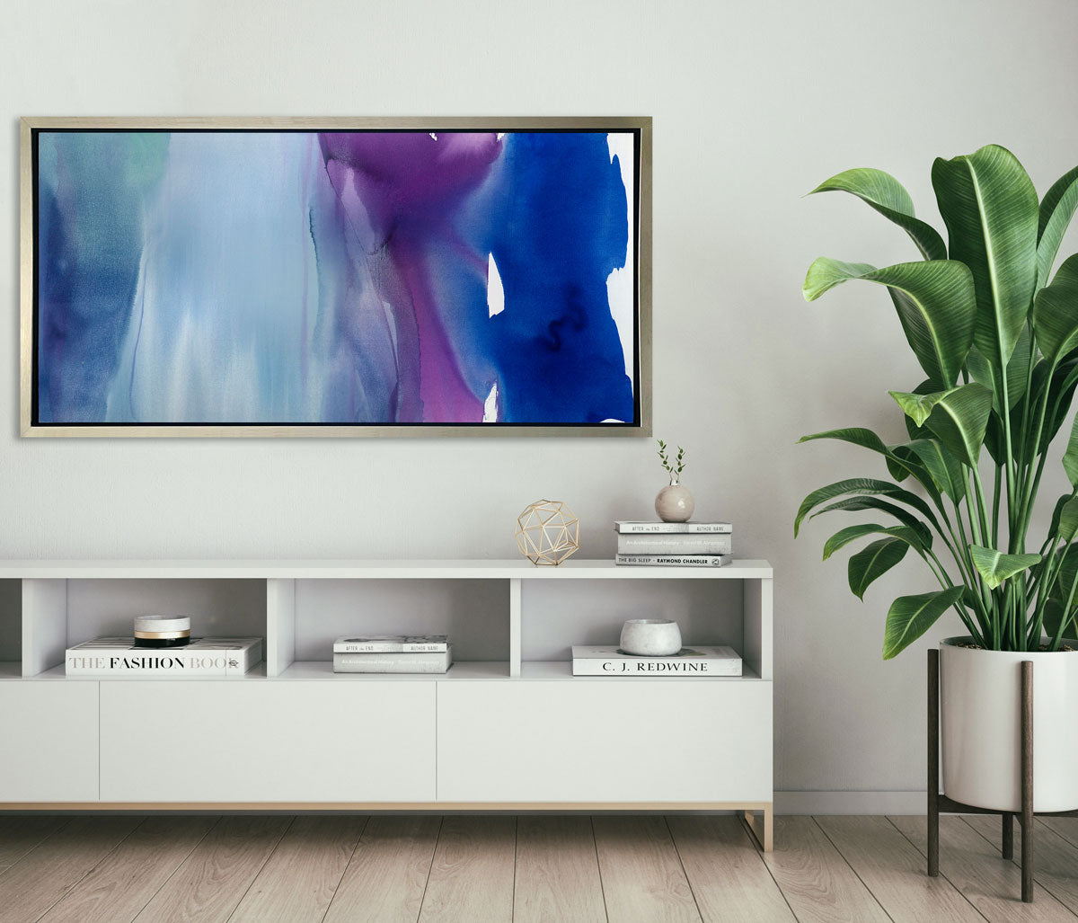 A blue, purple and light blue abstract print in a silver floater frame hangs on a light wall above a white credenza with stacks of books and a large potted floor plant to the right.