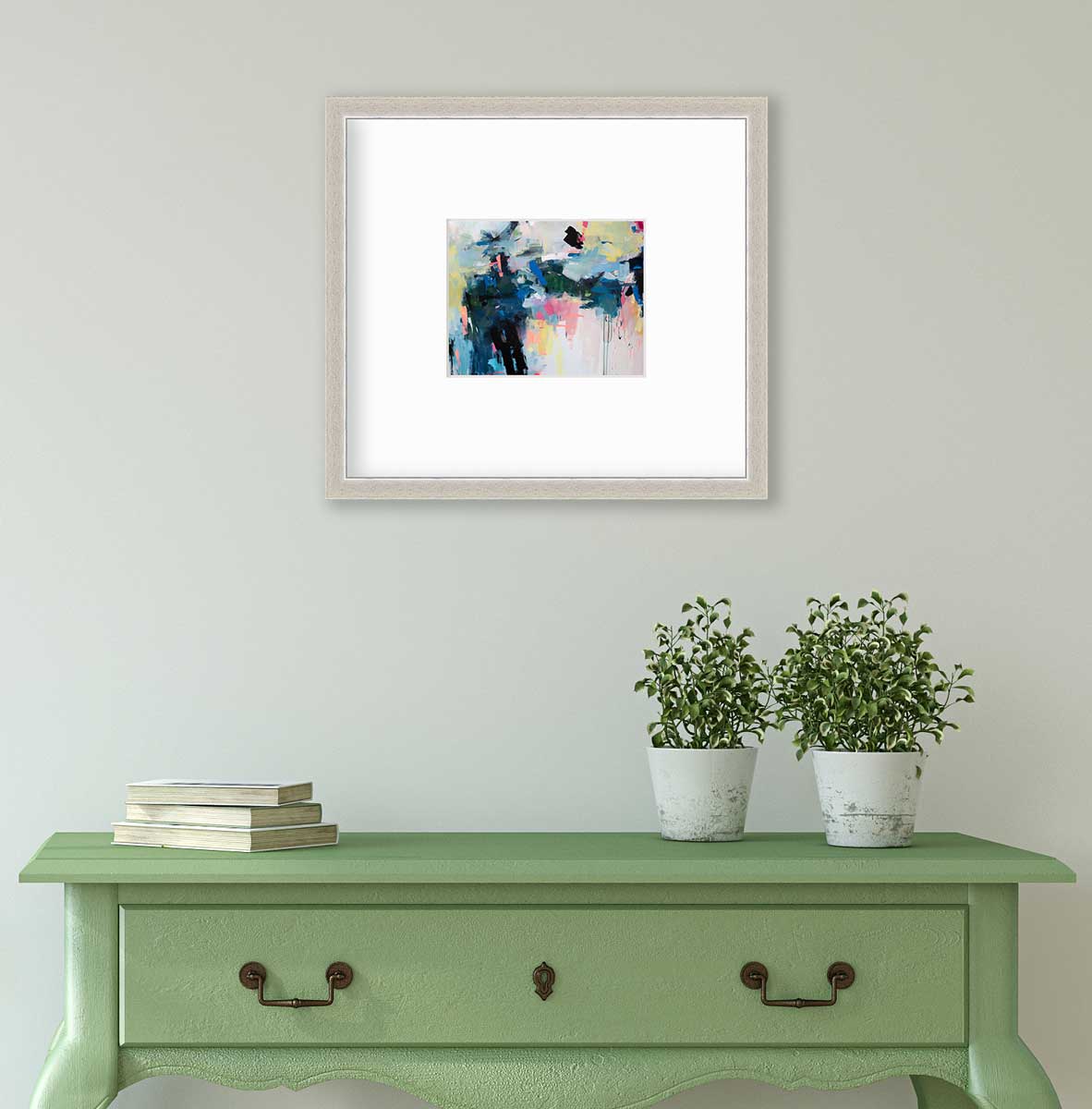 A colorful abstract print by Kelly Rossetti in a silver frame with a mat hangs on a white wall above a green console table with a stack of books and two potted plants.