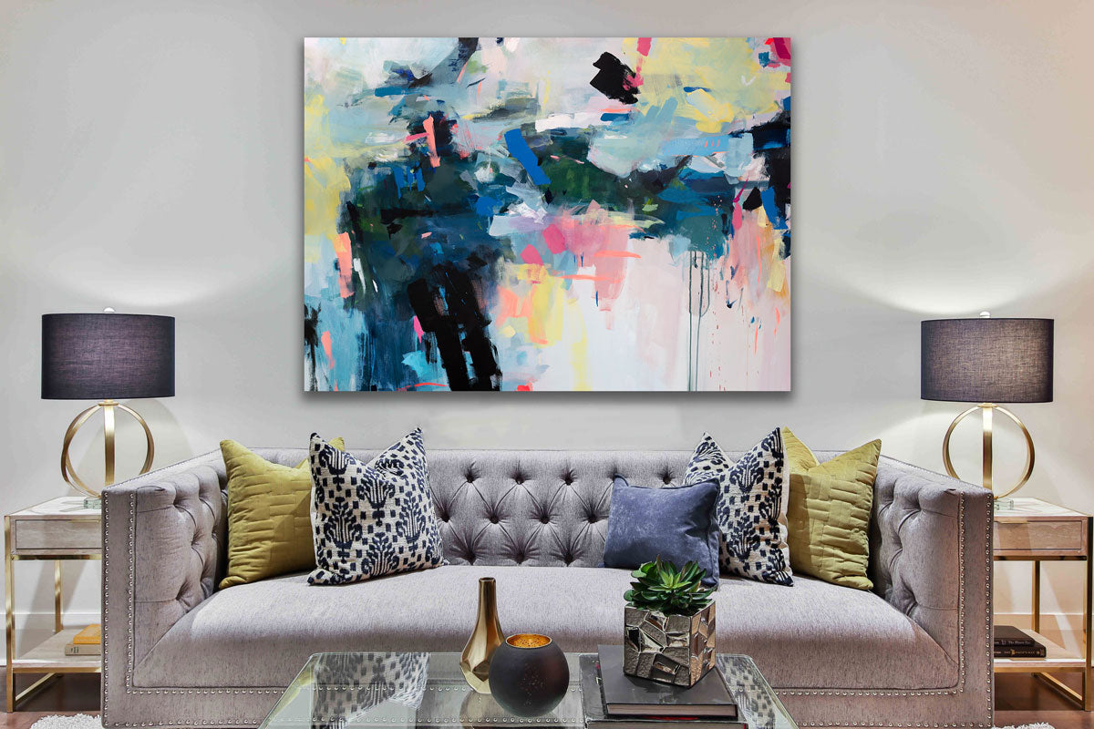 A colorful abstract painting hangs above a grey tufted sofa with pillows, two side tables with lamps and a glass rectangular coffee table with accessories.