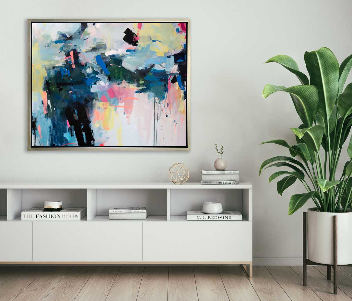 A multicolored abstract print in a silver floater frame hangs on a white wall above a white shelving unit with books, a gold geometric sphere and a small vase with greenery stems.