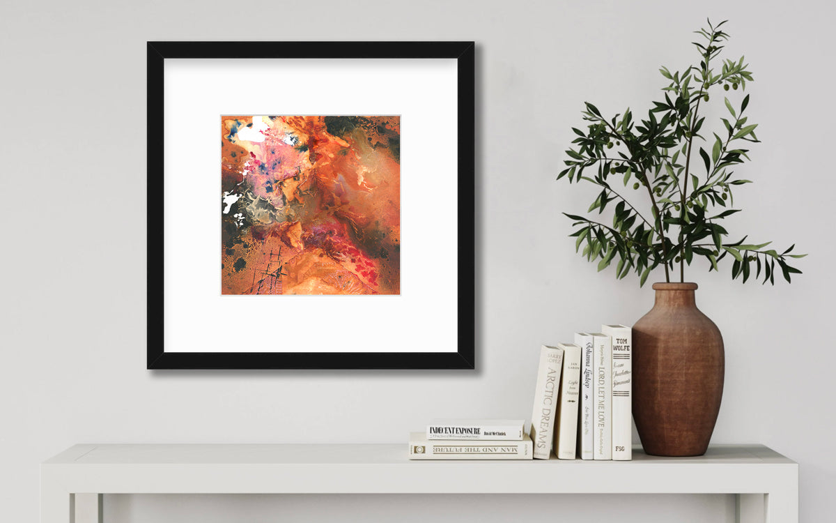 A red, orange, black and pink abstract print in a black frame with a mat hangs on a white wall above a white side table with a stack of books and a terracotta vase with greenery stems.