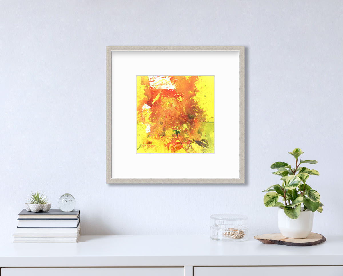 A yellow, orange and white abstract print in a silver frame with a mat hangs on a white wall above a white surface with a stack of books, a round clear container with gold accessories and a potted plant.