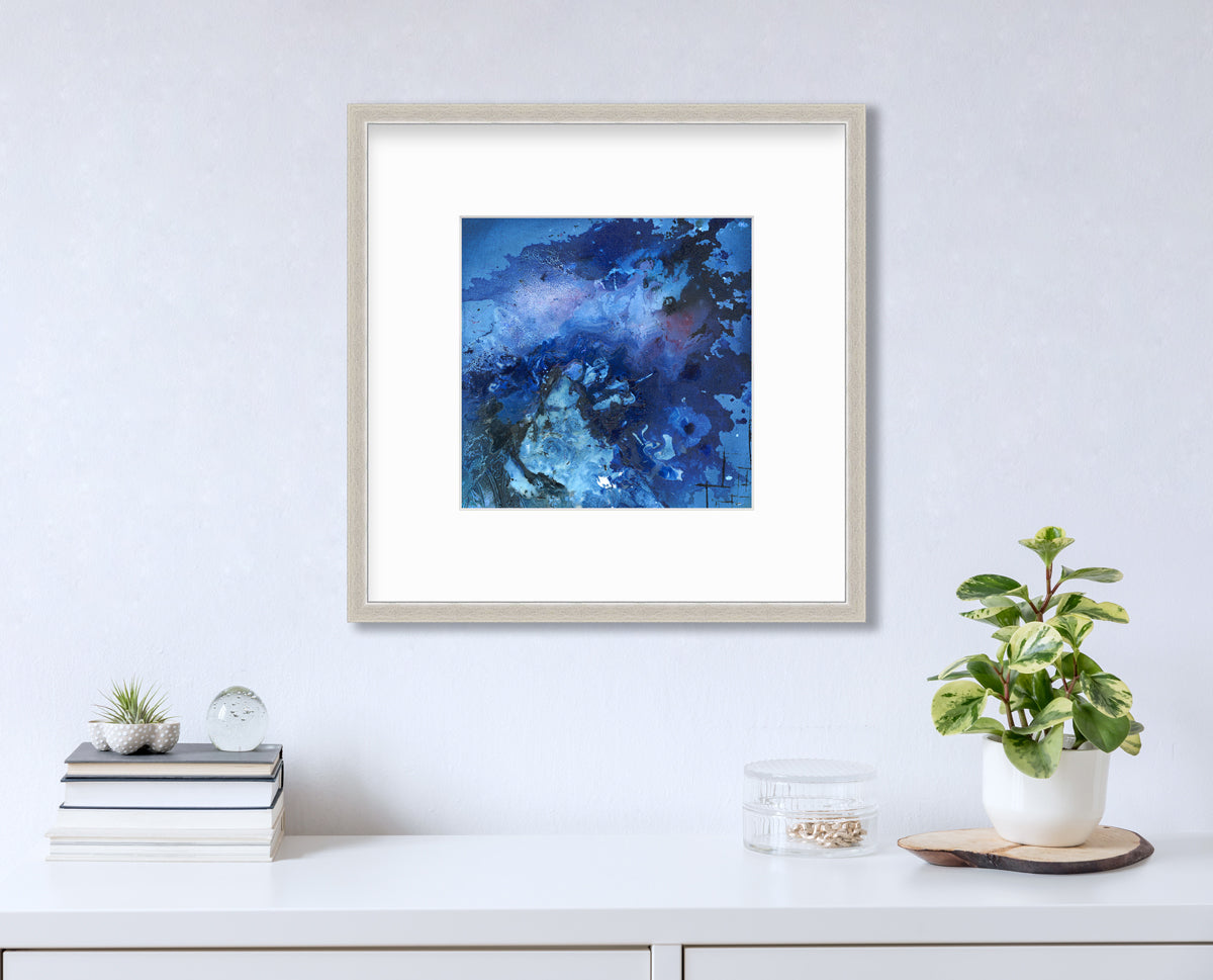 A blue and black abstract print in a silver frame with a mat hangs on a white wall above a white surface with a stack of books, a round clear container with gold accessories and a potted plant.