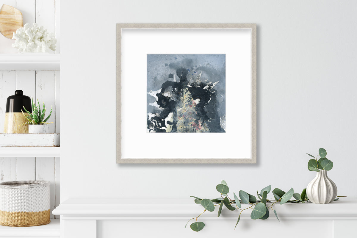 A grey, black, white and light yellow abstract print in a silver frame with a mat hangs on a white wall above a white shelf with a eucalyptus filled white vase and next to a built in book shelf.
