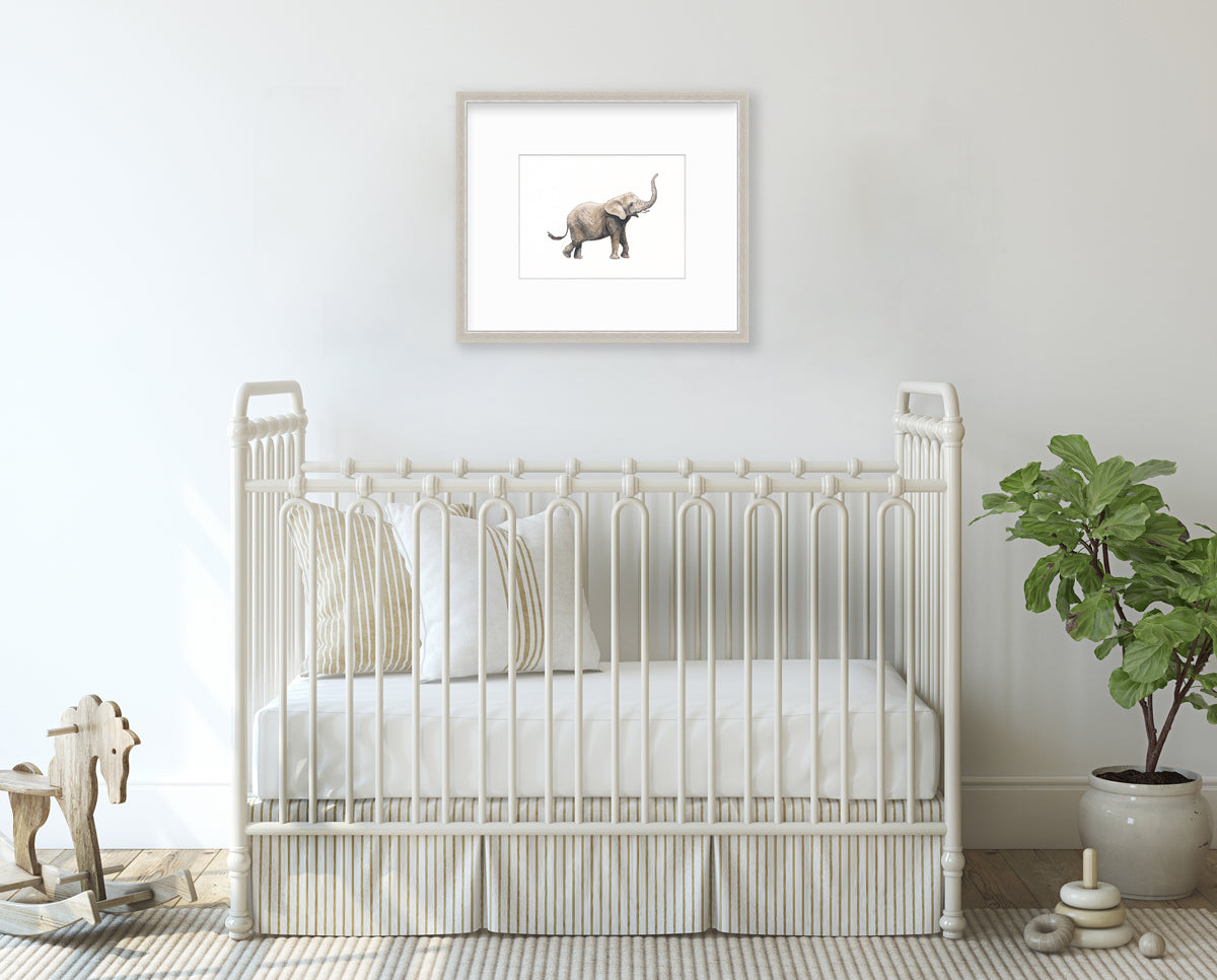 An elephant print in a silver frame with a mat hangs on a white wall above a white iron crib with neutral bedding, a wooden rocking horse and a potted floor plant.