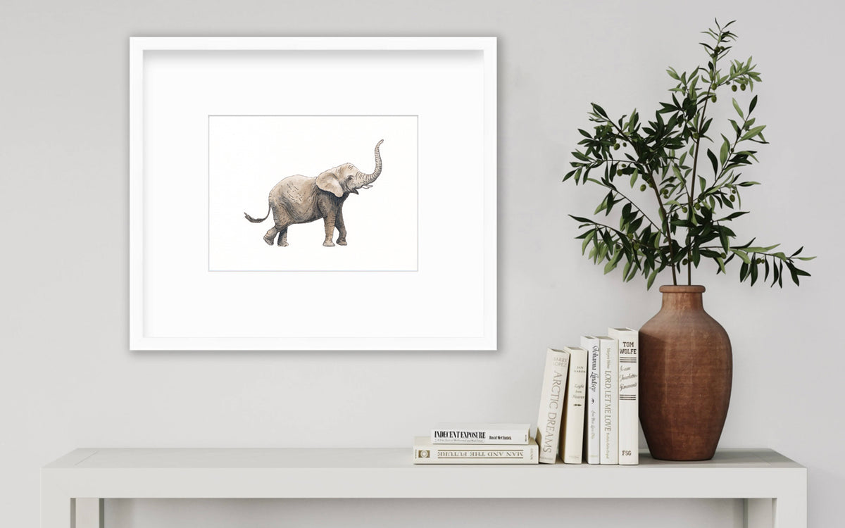 An elephant print in a white frame with a mat hangs on a white wall above a white surface with a stack of books and a terracotta vase with greenery stems.