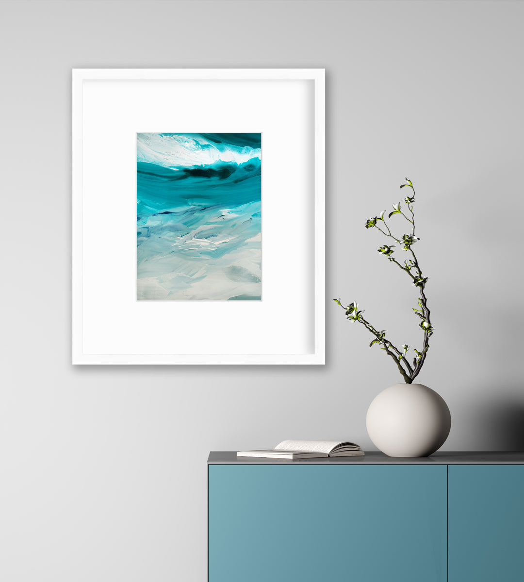 A teal, white and grey abstract print in a white frame with a mat hangs on a white wall above a teal cabinet with an open book and a white round vase with green stems.