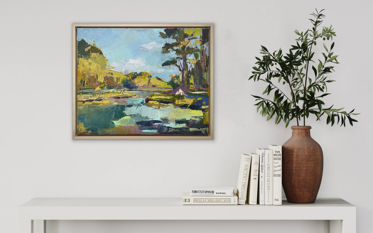 A blue and yellow abstract landscape painting framed in a natural floater frame hangs on a white wall over a white table with books and a pot holding green decorative branches.