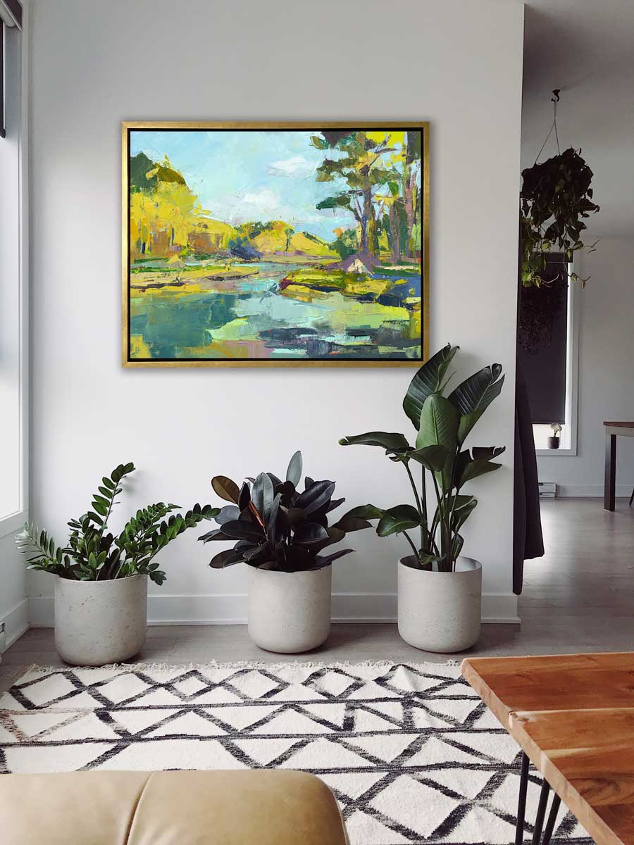 A chartreuse, light blue and green abstract landscape print in a gold floater frame hangs on a white wall above three various sized potted plants and a white and black printed rug.