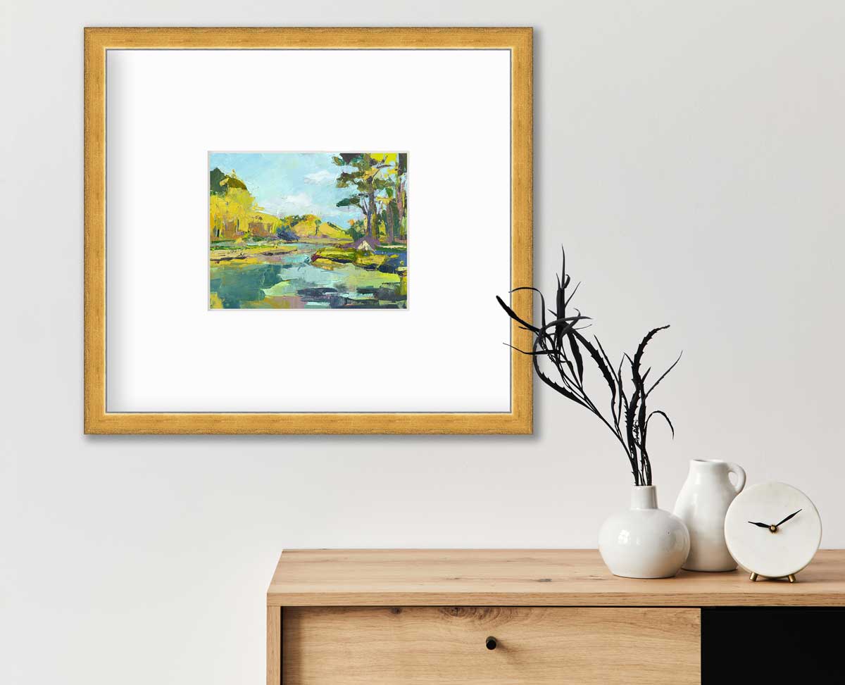 A chartreuse, light blue and green abstract landscape print in a gold frame with a mat hangs on a white wall above a wood side table with a white modern clock, a small white pitcher and a white vase with dark green leaves.