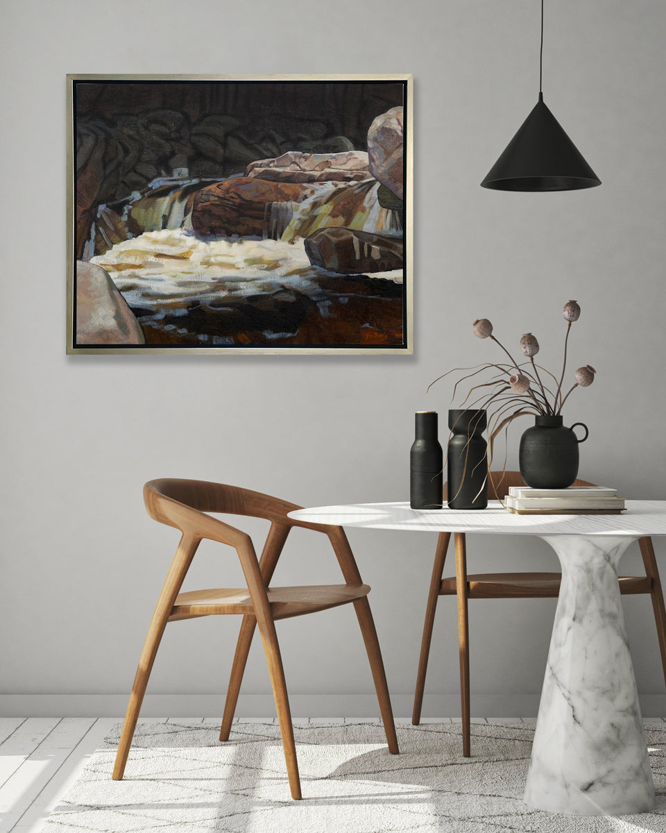 A brown, black and white abstract river landscape print in a silver floater frame hangs on a white wall above a round white marble pedestal table with black vases and two wood chairs.