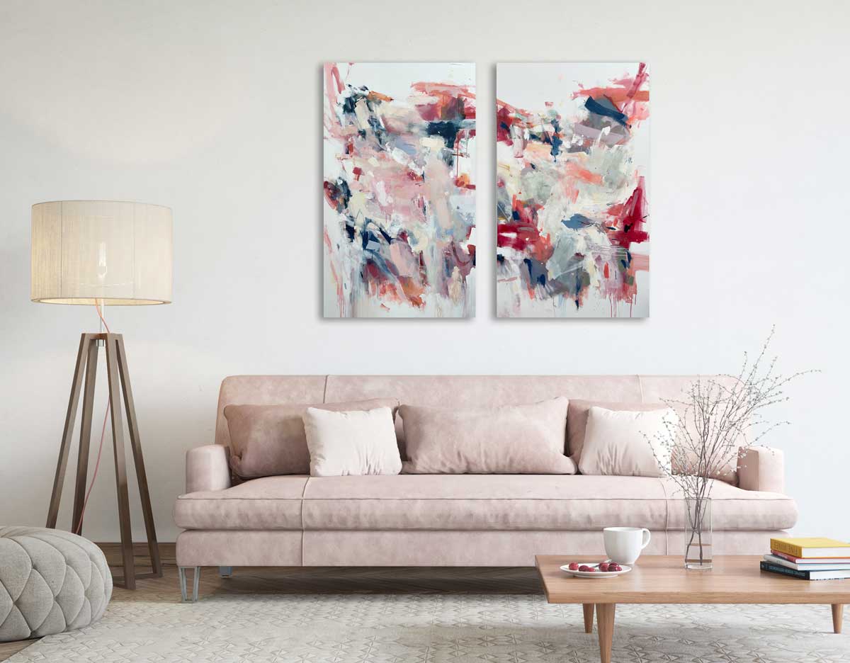 Two light pink and white abstract paintings by Kelly Rossetti hang on a white wall above a light pink sofa with pillows, a low wooden square coffee table and a four polled floor lamp.