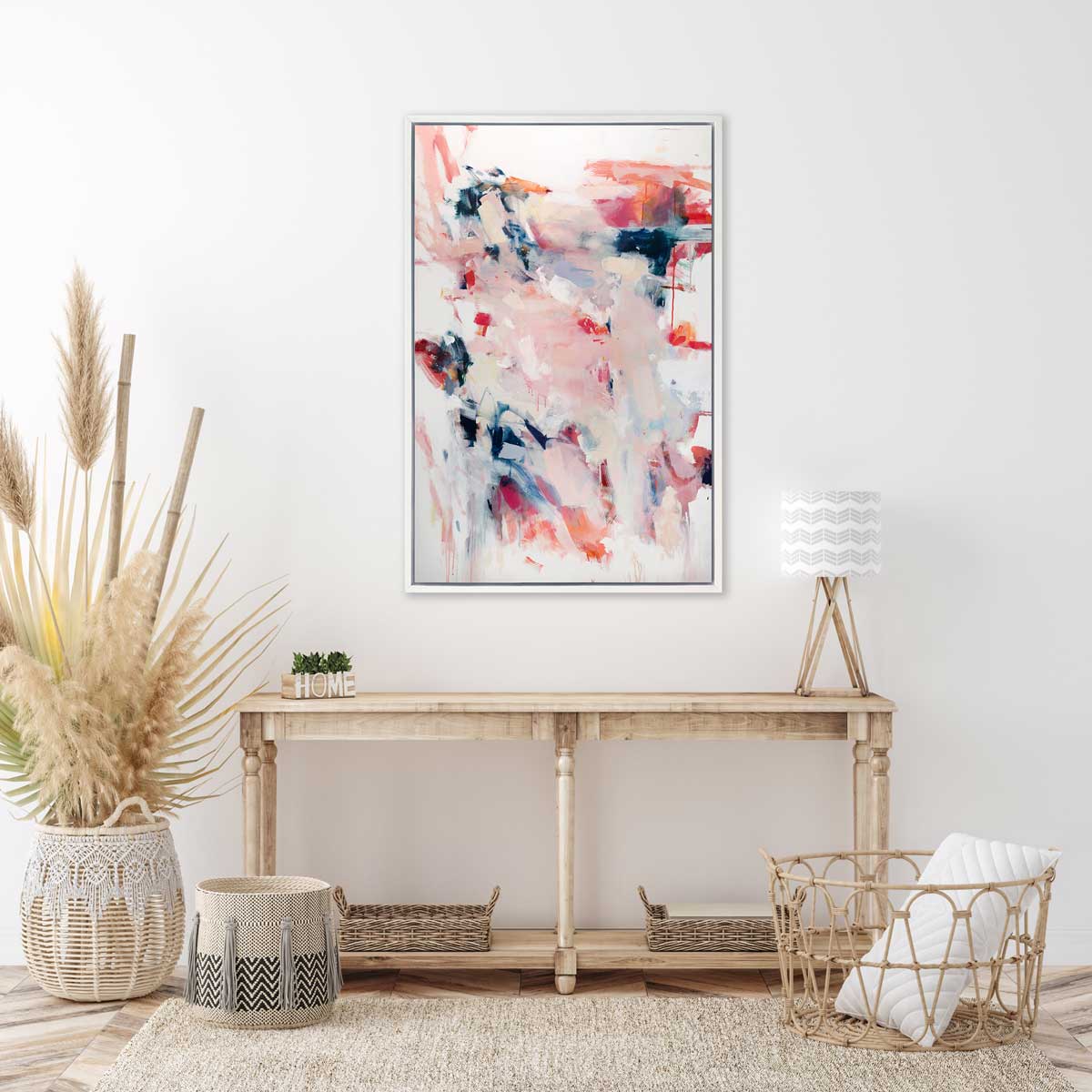 A pink, white and blue abstract print in a white floater frame hangs on a white wall above a natural wood console table with a small table lamp, several woven baskets and a small decorative wood vase with greenery.