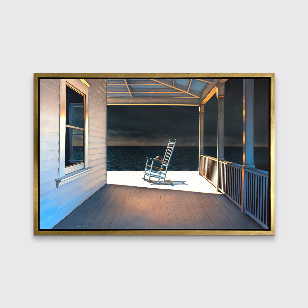 A print of a white rocking chair on a deck of a house with a high contrast light on the chair in a gold floater frame hangs on a white wall.