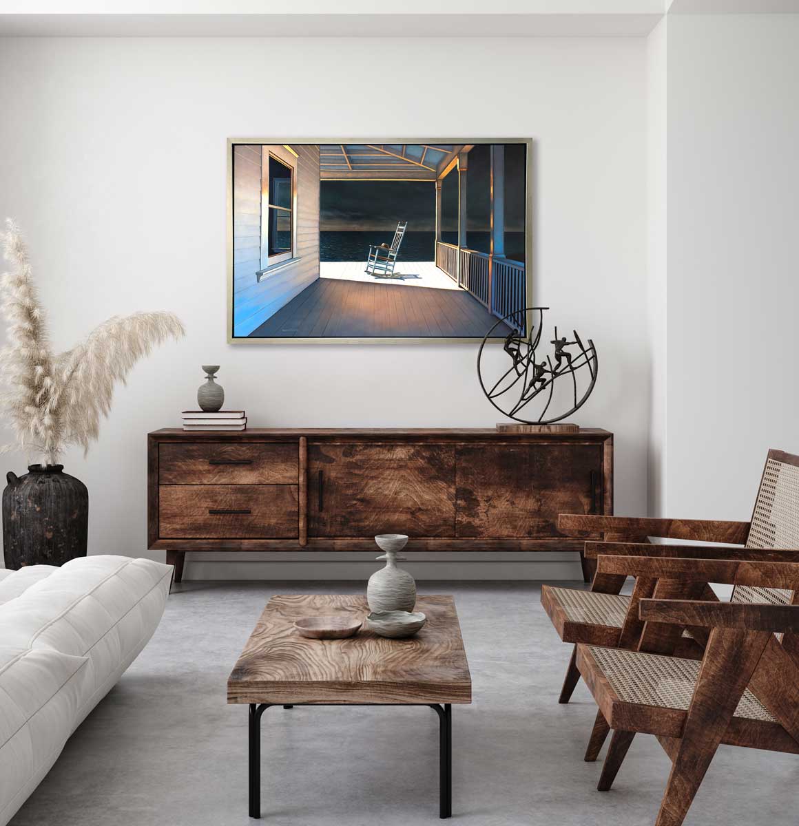 A print of a white rocking chair on a deck of a house with a high contrast light on the chair in a silver floater frame hangs on a white wall above a stained wood console stand with a black decorative accessory, a small vase under stacked books, two stained wood chairs and a wood coffee table.
