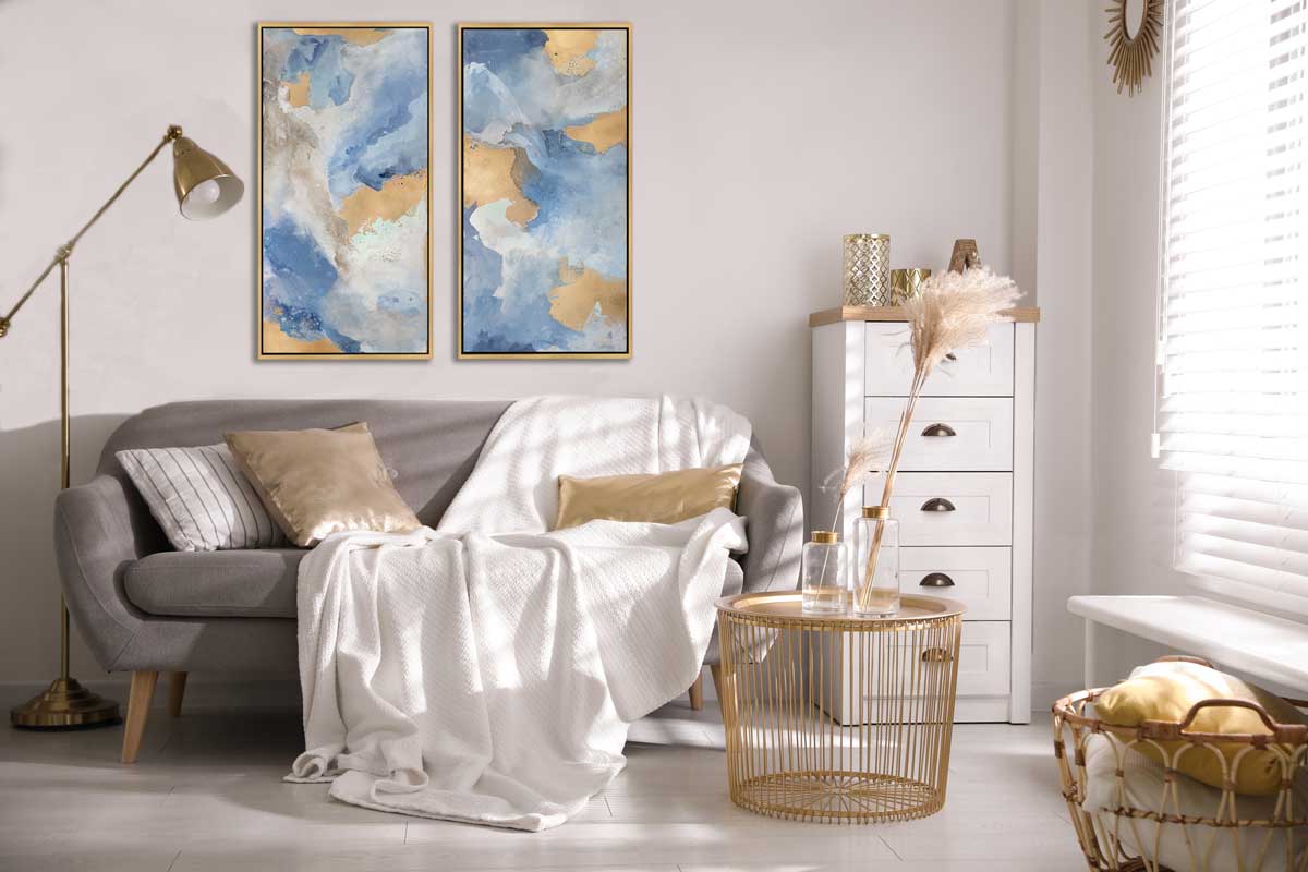 Two blue, gold and white abstract prints in gold floater frames hang on a white wall above a grey sofa with a large white blanket draped over, a round gold coffee table and a tall white filing cabinet.