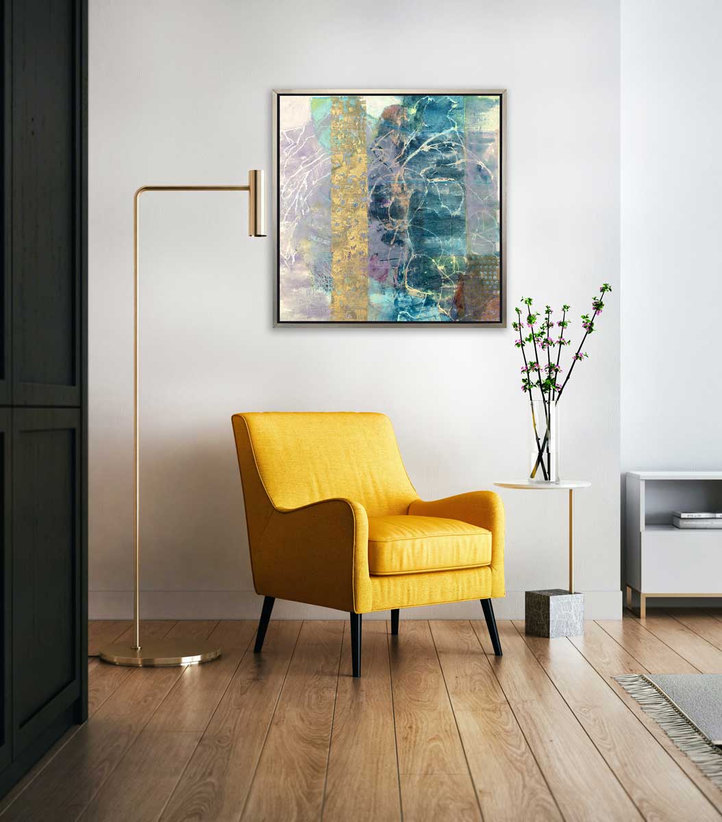 A blue, purple and gold abstract print in a silver floater frame hangs on a white wall above a yellow chair, a side table with a vase of purple flowers and a large gold floor lamp.