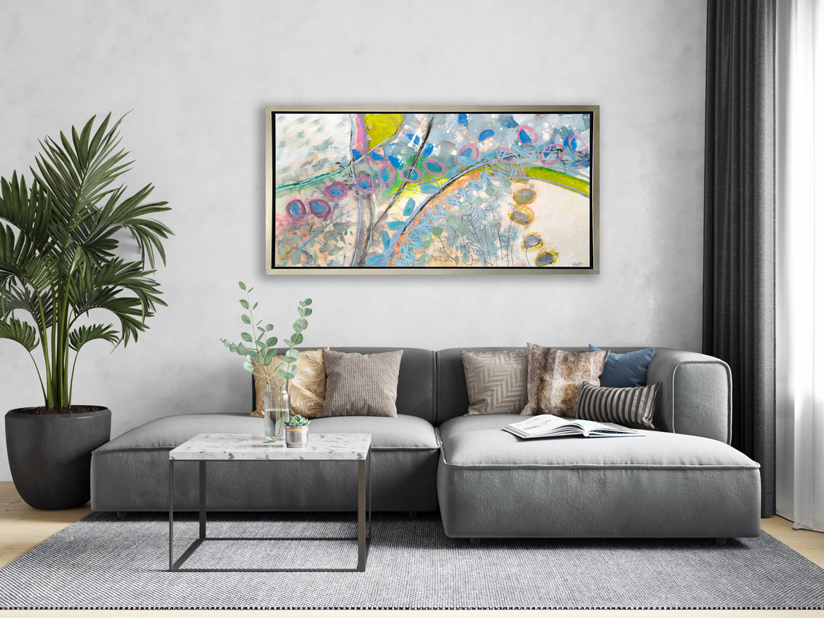 A multicolored abstract print in a silver floater frame hangs on a grey wall above a oversized grey sectional with pillows, a square white marble coffee table and a tall potted floor plant.