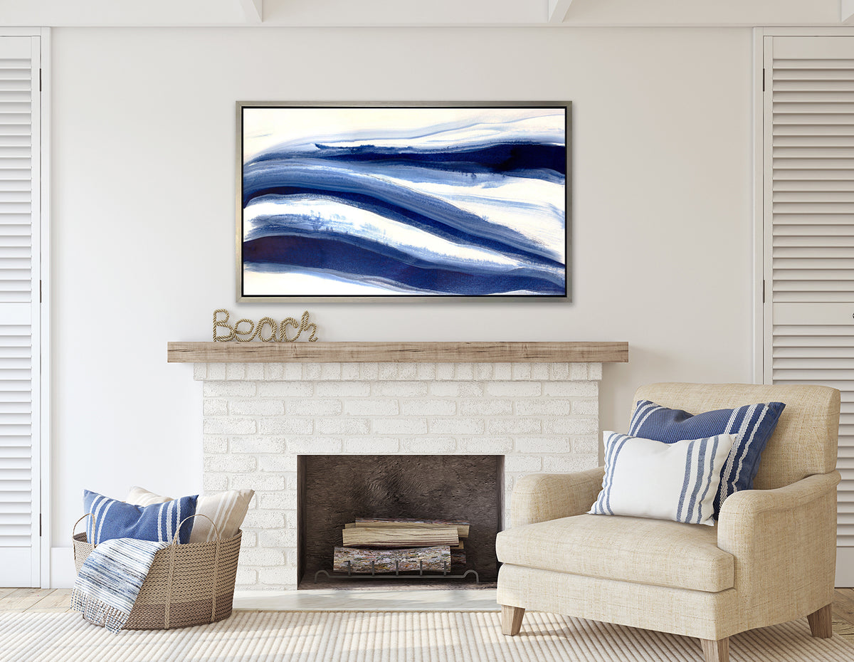 A dark blue and white abstract print in a silver floater frame hangs on a white wall above a white brick fireplace, a cream linen chair with pillows and a woven basket with pillows.