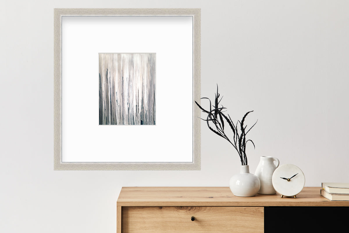 A white, cream and charcoal print in a silver frame with a mat hangs on a white wall above a wood console table with a white vase with stems, a small white pitcher and a white modern clock.