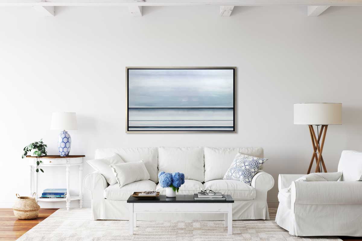 A tonal blue abstract landscape print in a silver floater frame hangs on a white wall above a white sofa with pillows, a matching overstuffed chair, a black and white coffee table with accessories, a small side table with a blue and white table lamp and a tall floor lamp.