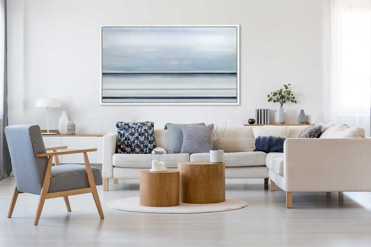A tonal blue abstract landscape print in a white floater frame hangs on a white wall above a white sectional sofa with blue, grey and white pillows, a blue and white checkered chair and two round solid wood tables in the center.