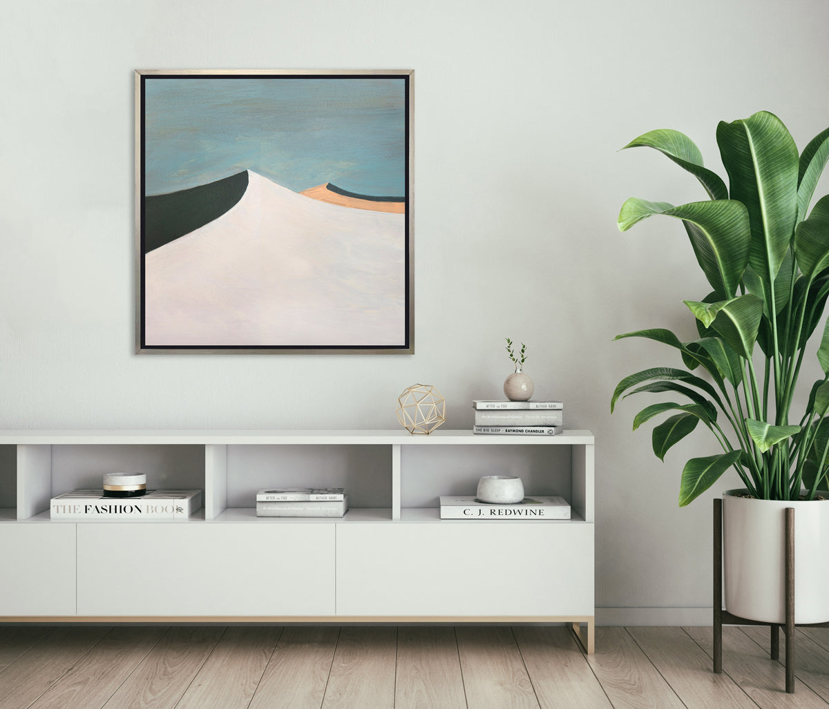 A blue, white, peach and dark teal abstract landscape print in a silver floater frame hangs on a white wall above a white shelf console with books, a gold wire sphere decoration and a large potted floor plant to the right.