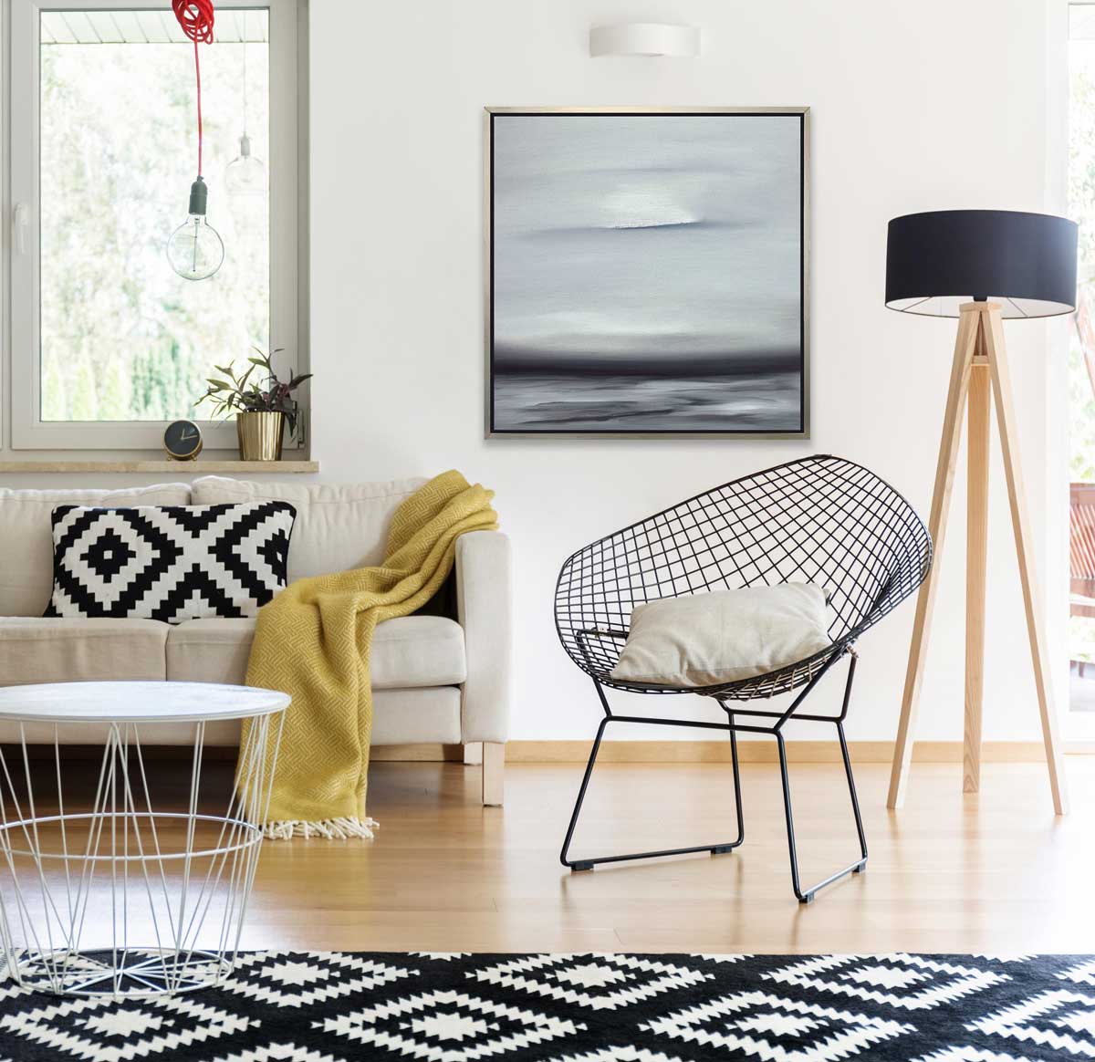 A tonal grey and black print in a silver floater frame hangs on a white wall above a black wire chair, a tripod floor lamp and a white sofa with a black and white pillow and a yellow throw blanket.