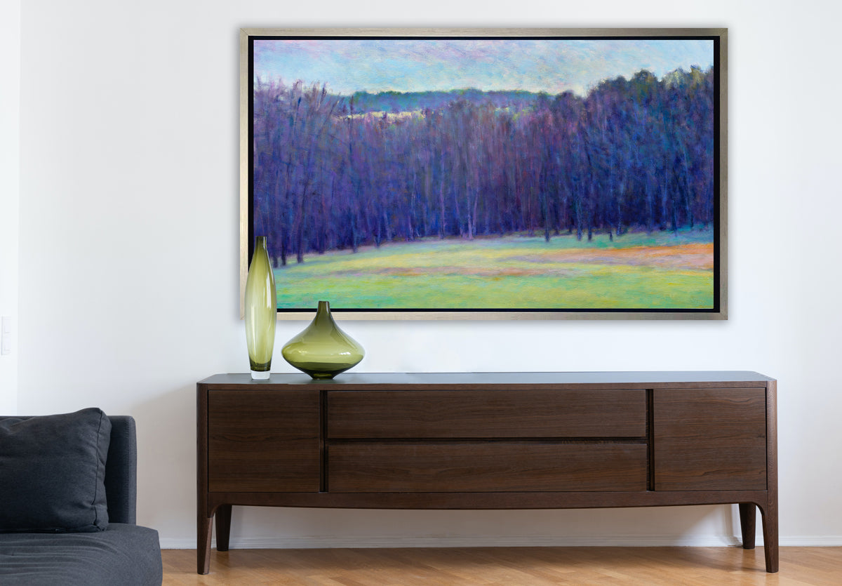 A blue, purple, light green and light orange impressionistic landscape print in a silver floater frame hangs on a white wall above a stained wood credenza with two green glass vases and near the side of a denim blue sofa.