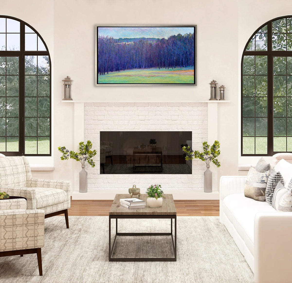 A blue, purple, light green and light orange impressionistic landscape print in a silver floater frame hangs on a white wall above a white brick fireplace, two white and grey printed armchairs, a white sofa with pillows and a natural wood coffee table sits center with decorative accents.