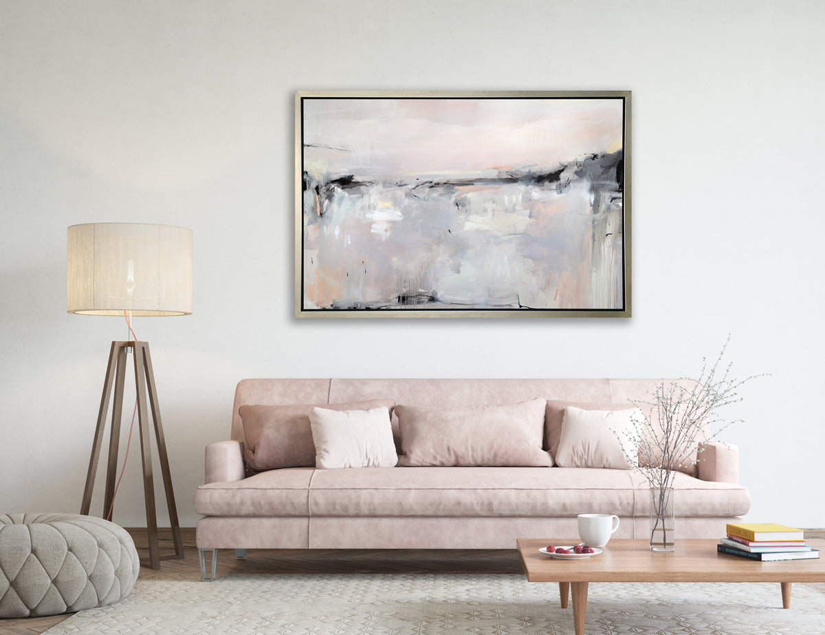 A light pink, black, grey and white abstract print in a silver floater frame hangs on a white wall above a light pink sofa with pillows, a low wood coffee table with decorative items, a large floor lamp and a grey tufted ottoman.