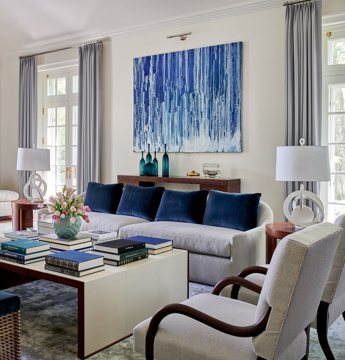 A blue and white abstract drip painting by Teodora Guererra hangs on a white wall in a formal living room. The painting is hung above an espresso accent table holding teal vases and a glass bowl. In front of the console table is a grey modern couch with blue throw pillows and a beige coffee table in front holding various stacked books. On either side of the couch are wooden side tables with white modern lamps. 