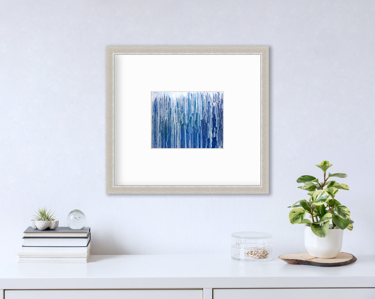 A blue and white abstract print in a white frame with a mat hangs on a white wall above a white surface with a stack of books, a round clear container with gold accessories and a potted plant.