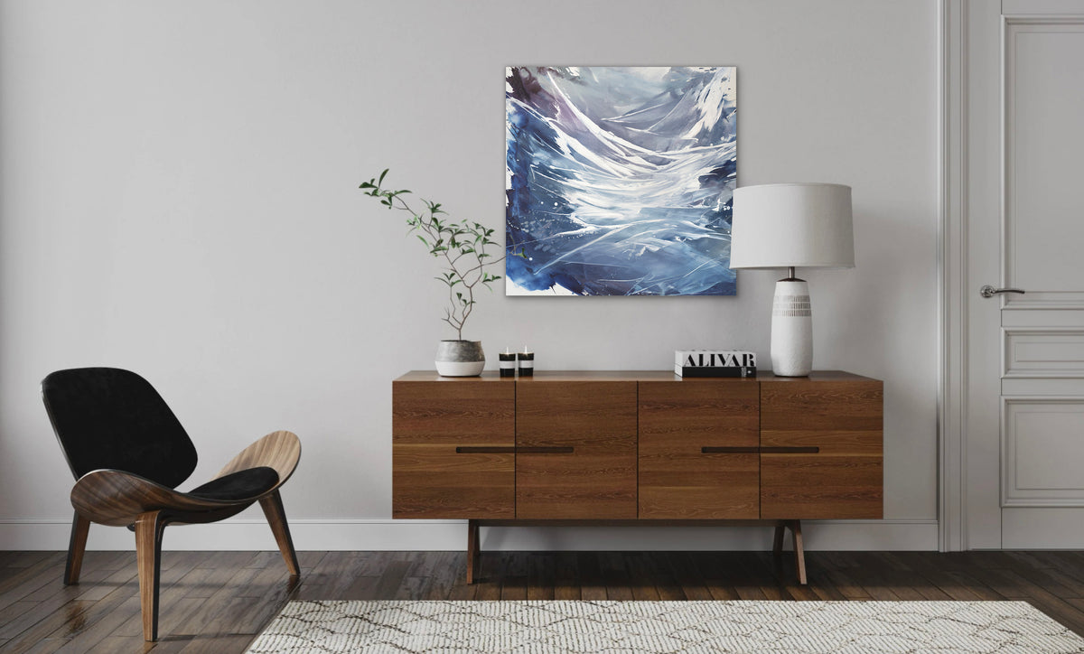 A blue, white, and purple abstract painting by Teodora Guererra hangs on a white wall above a modern wood console. On top of the console sits a white lamp, coffee table books, and a potted plant. To the left of the console table is a modern wooden chair.