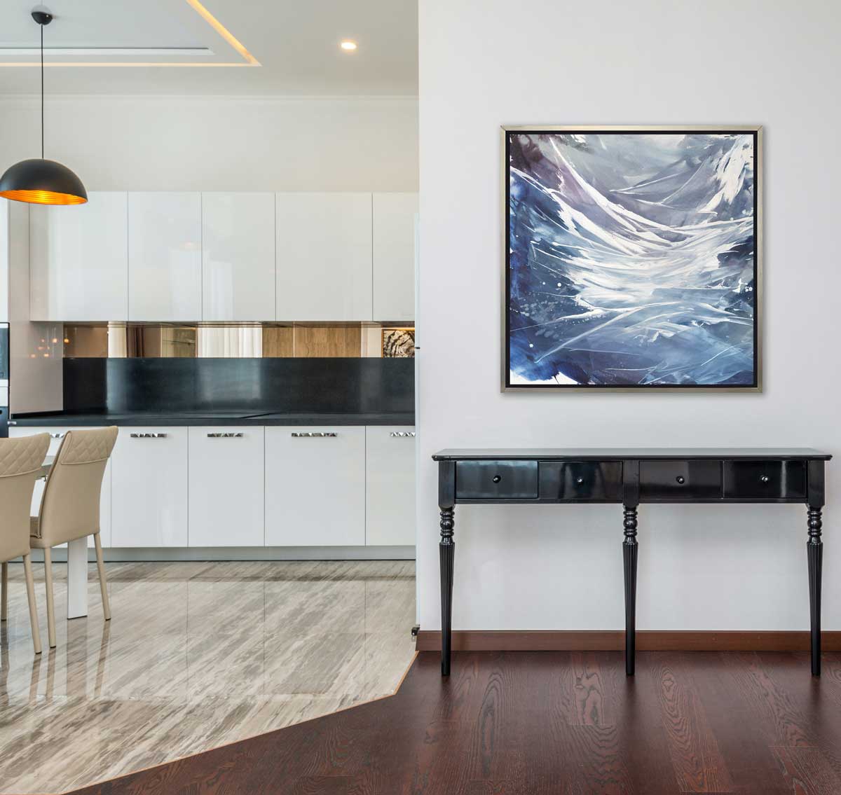 A light purple, grey, white and blue abstract print in a silver floater frame hangs on a white wall above a black console table and next to an opening to a modern kitchen with white cabinets, black countertops and two beige counter stools.