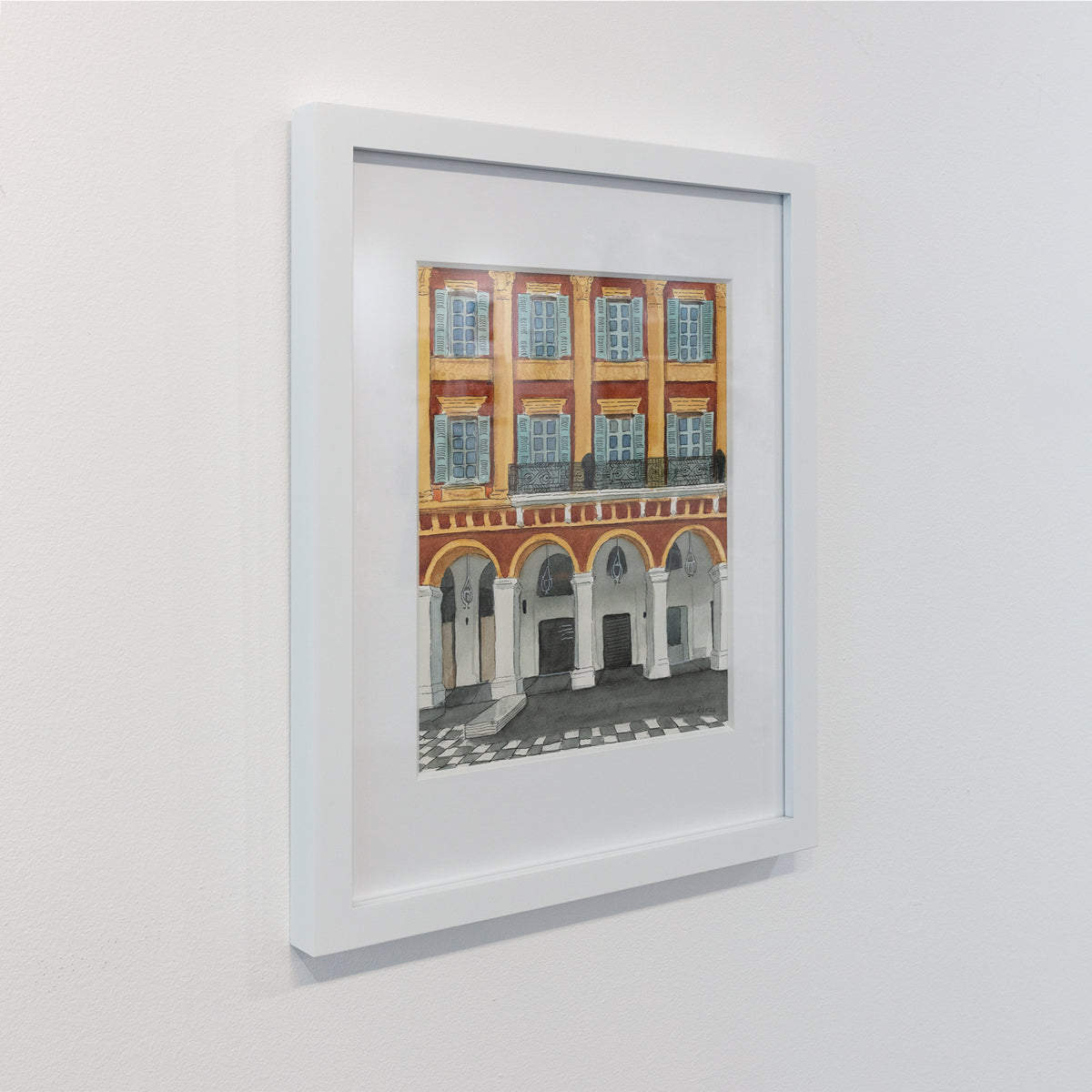 A side-angle view of an orange and yellow architectural painting in a white frame, hanging on a white wall. 