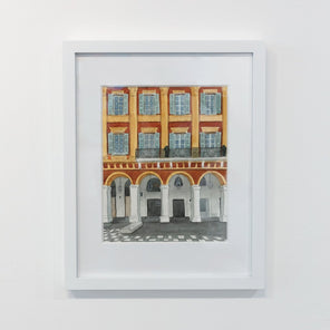 A painting of orange and yellow architecture, framed in a white frame, hangs on a white wall. 