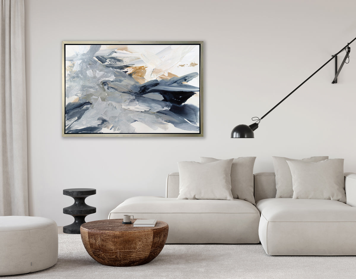 A grey, blue, gold and white abstract print in a silver floater frame hangs on a white wall above a white sectional sofa with pillows, a rounded wood coffee table and a black movable lamp attached to the wall.
