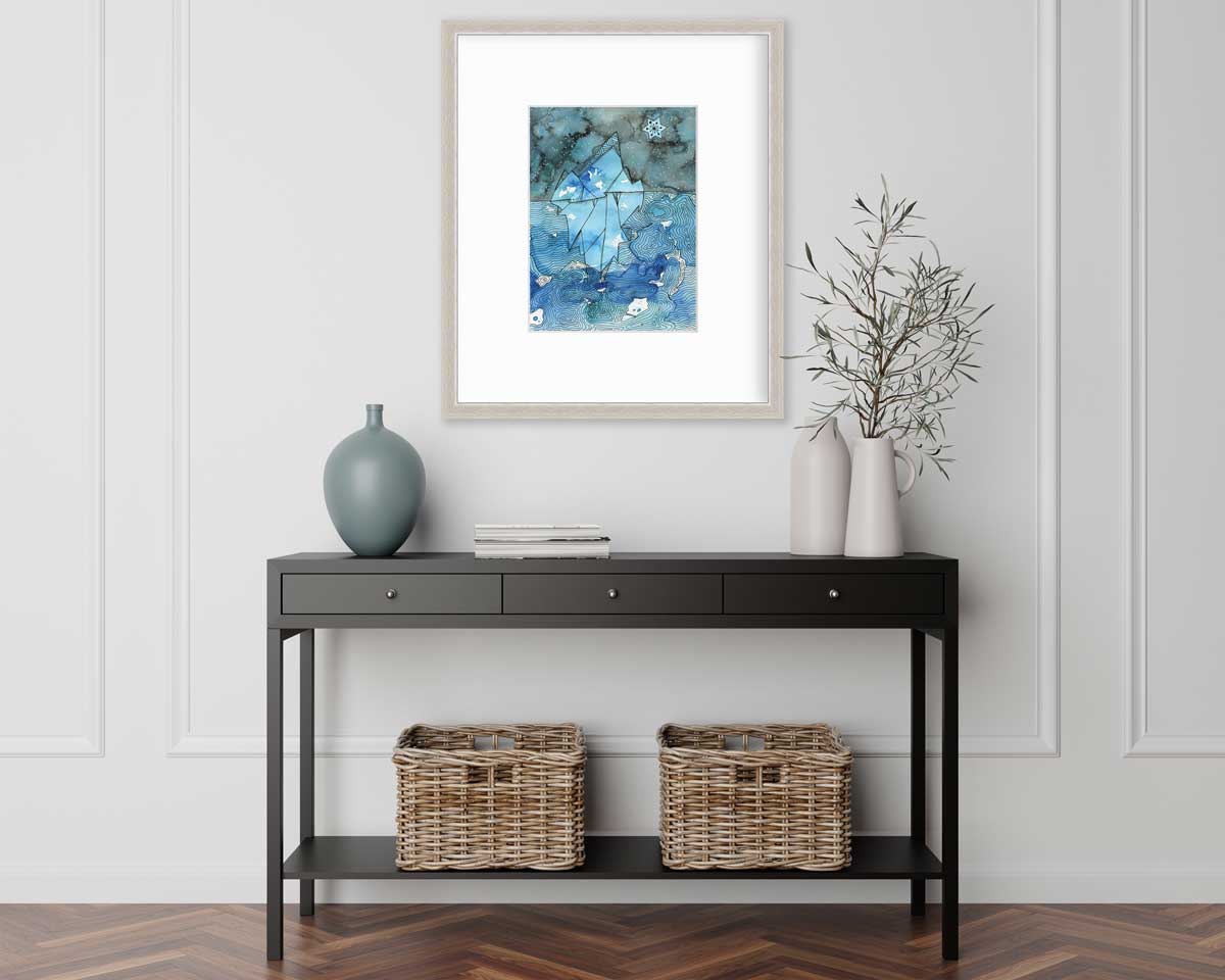 A blue abstract glacier illustration print in a silver frame with a mat hangs on a white wall above a black console table with a muted teal vase, two white vases with greenery stems and two wicker baskets.