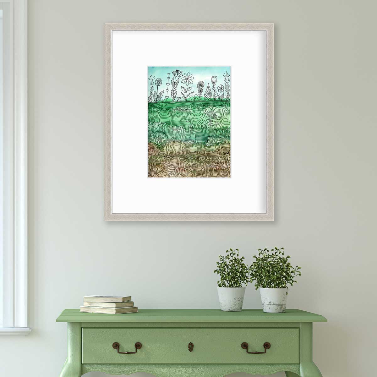 A green, light blue and brown botanical illustration print in a silver frame with a mat hangs on a white wall above a green console table with a stack of books and two potted plants.