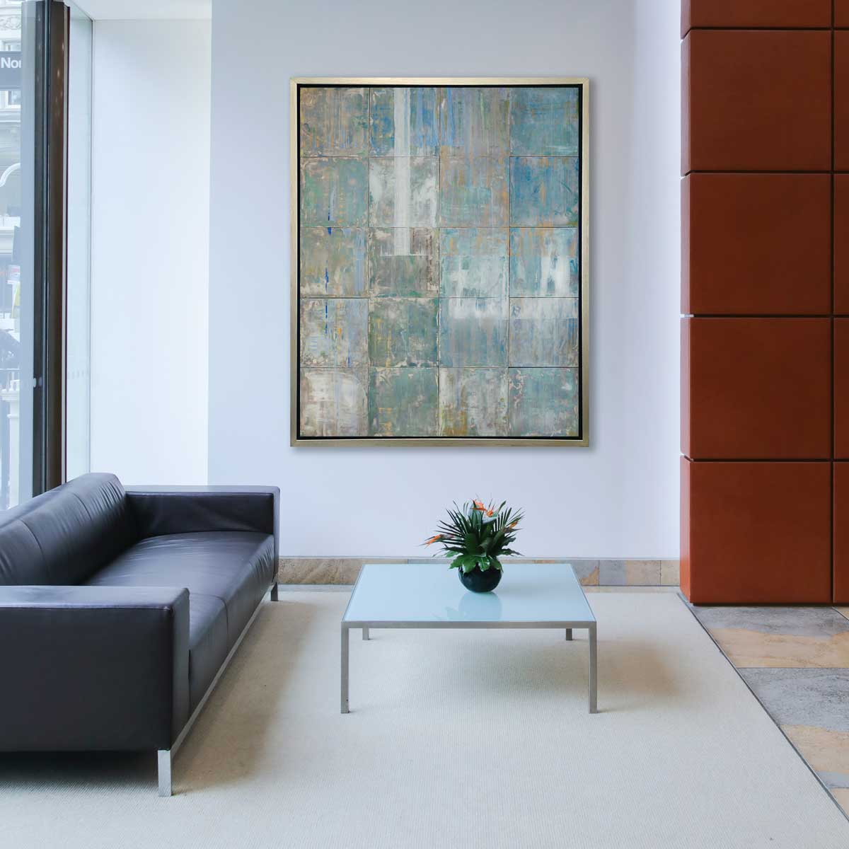 A blue and green abstract geometric print in a silver floater frame hangs on a white wall across from a black leather sofa and a small square frosted glass coffee table.