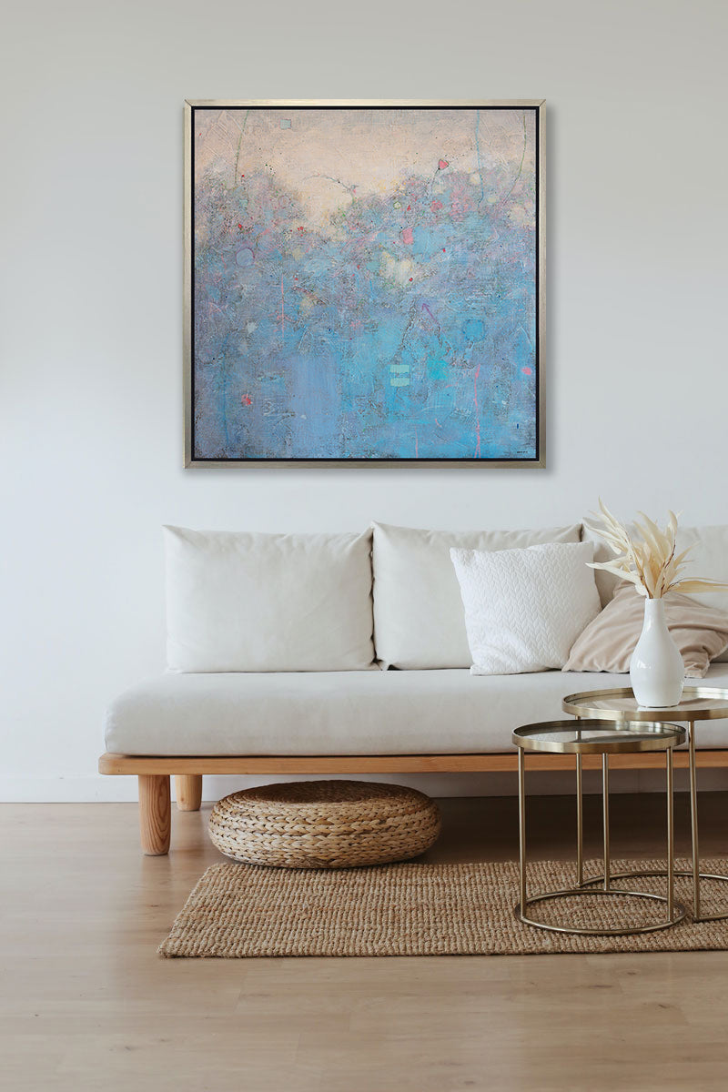 A blue abstract landscape print by Elwood Howell in a silver floater frame hangs on a white wall above a wooden plank couch with white cushions, a woven ottoman and a two tier coffee table.