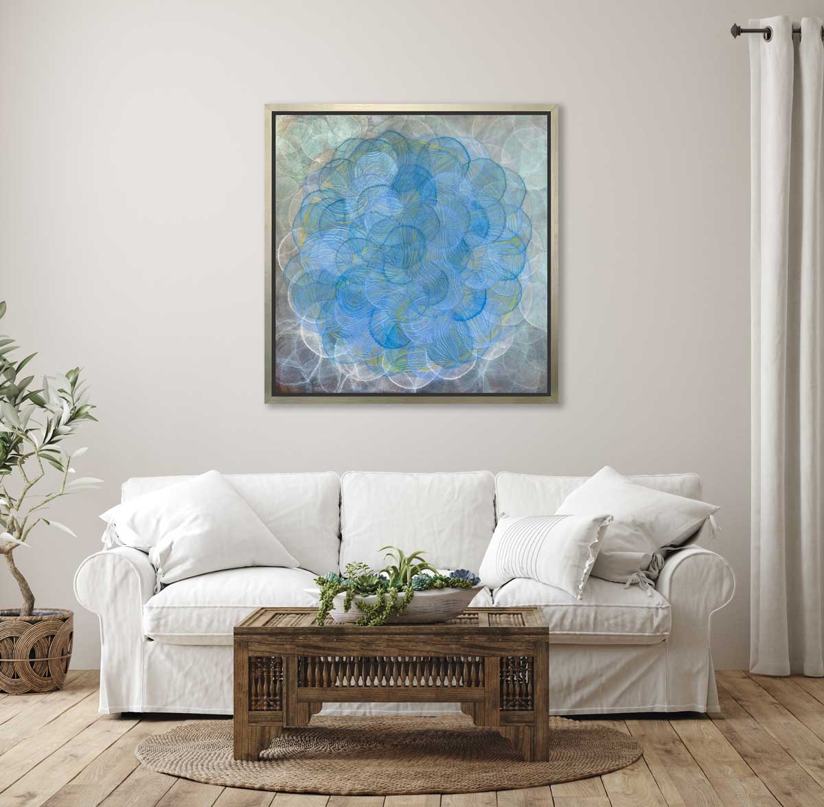 A blue, white and teal overlapping circles print in a silver floater frame hangs on a white wall above a white sofa with white pillows and a ornate wood rectangular coffee table with a bowl of green and blue succulents.