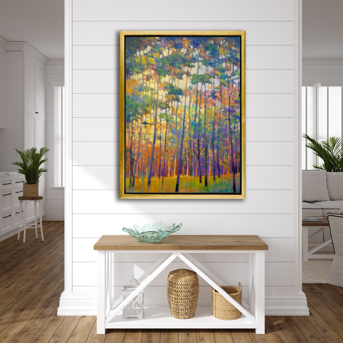A multicolored abstract tree landscape print in a gold floater frame hangs on a white shiplap wall above a butcher block and white wood console table with a teal green glass bowl, two woven baskets and a white lantern.