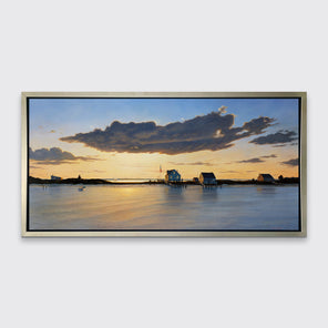 A print of three bay houses on a strip of land at sunset in a silver floater frame hangs on a white wall.