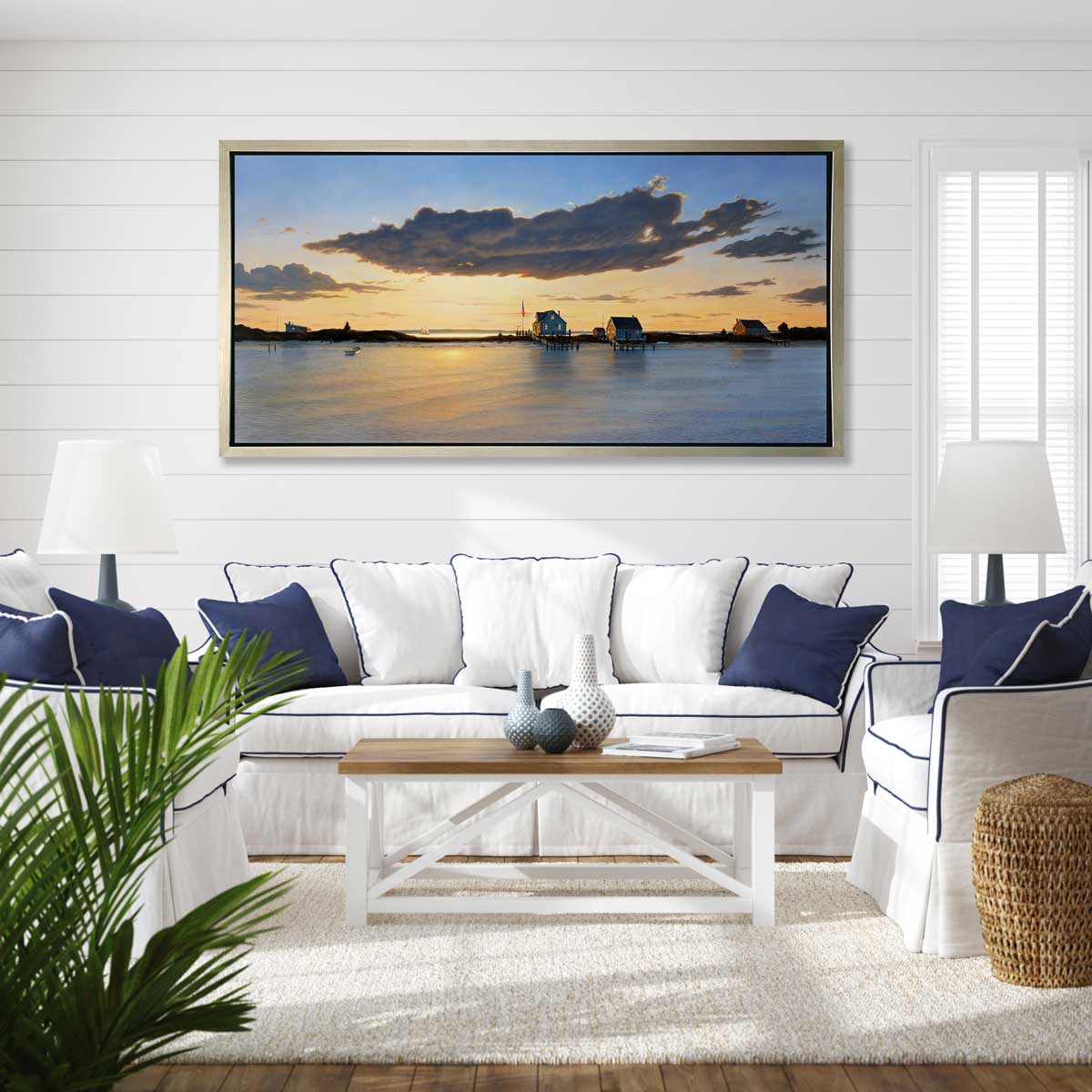 A print of three bay houses on a strip of land at sunset in a silver floater frame hangs on a white wall above a white sofa with white and blue pillows with matching white chairs and a coffee table with vases and books.