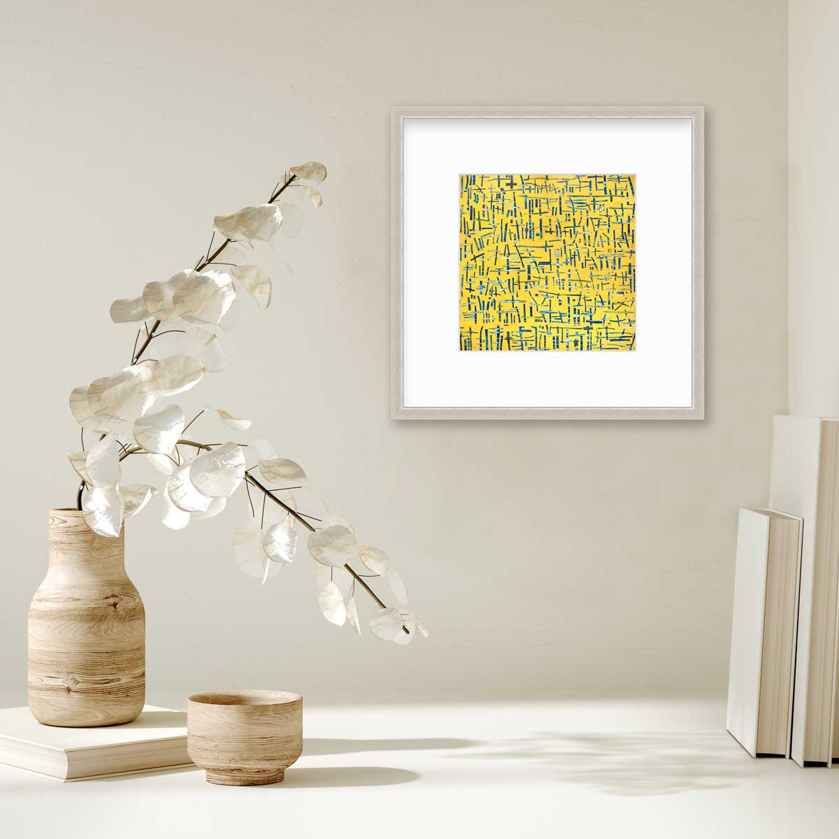 A yellow, blue and white abstract print in a silver frame with a mat hangs on a white wall above a stack of books and a wooden vase with white leaf stems.