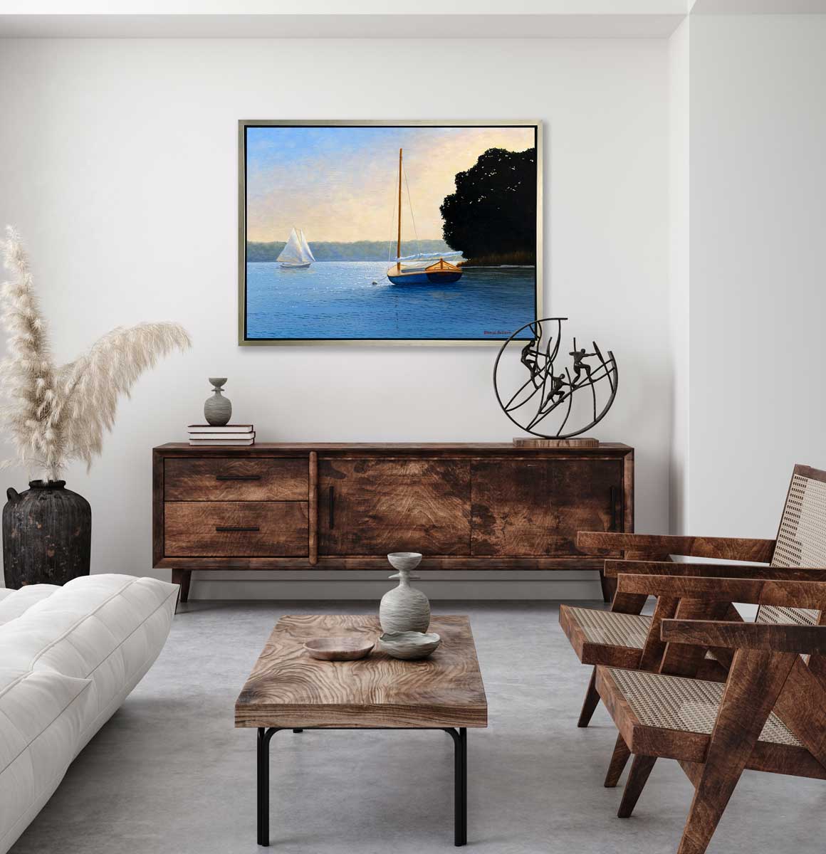 A blue, beige and black contemporary seascape print with two sailboats in a silver floater frame hangs on a white wall above a stained wood sideboard with books, a sculpture and stained wood chairs.