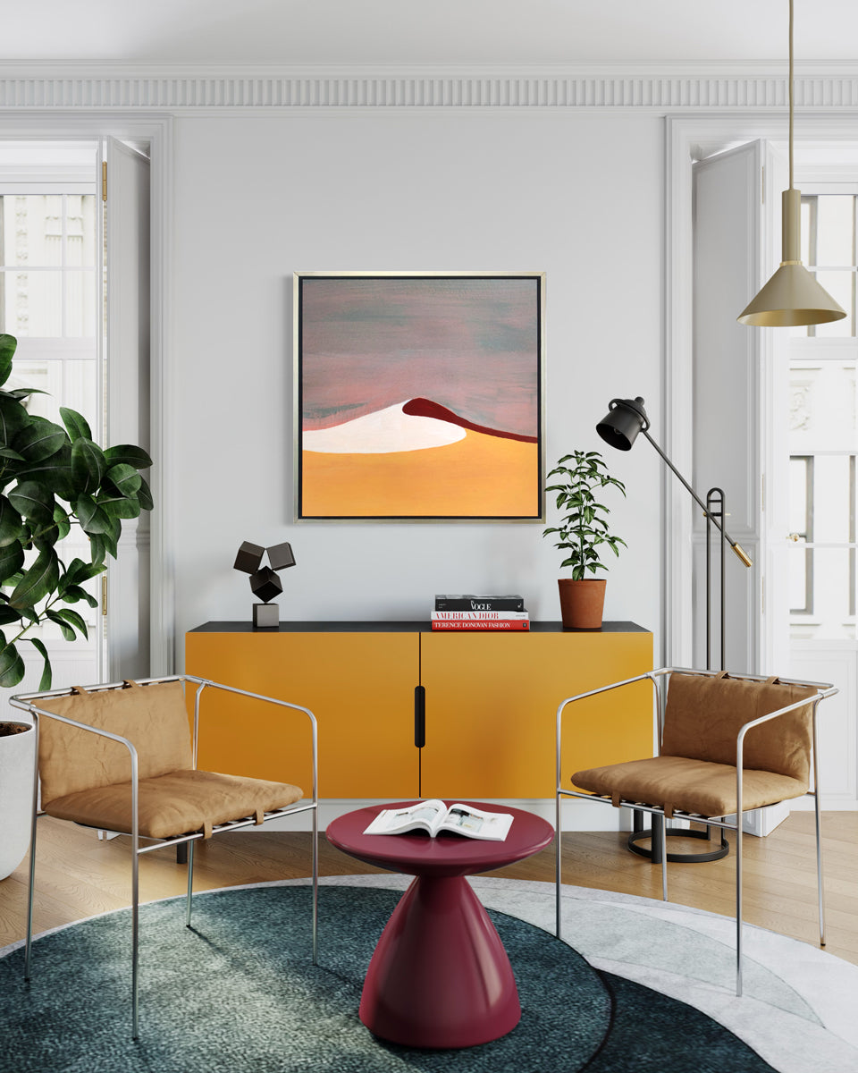 A mauve, burnt yellow, red and white abstract landscape print in a silver floater frame hangs on a white wall above a dark yellow credenza with accessories, two chrome armchairs with beige cushions and a raspberry pedestal side table.